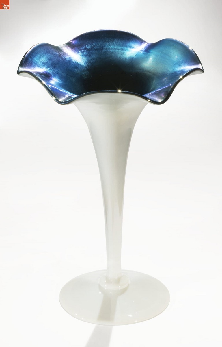 White glass vase with stem widening out from narrow at bottom to a crenellated flower shape opening at top; inside is iridescent blue