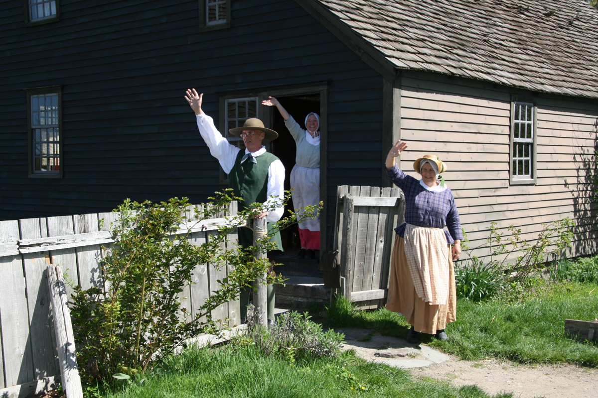 Presenters Working at Daggett Farmhouse in Greenfield Village, April 2006 Three people in historic garb wave from the doorway and yard of a gray wooden building with a wooden fence