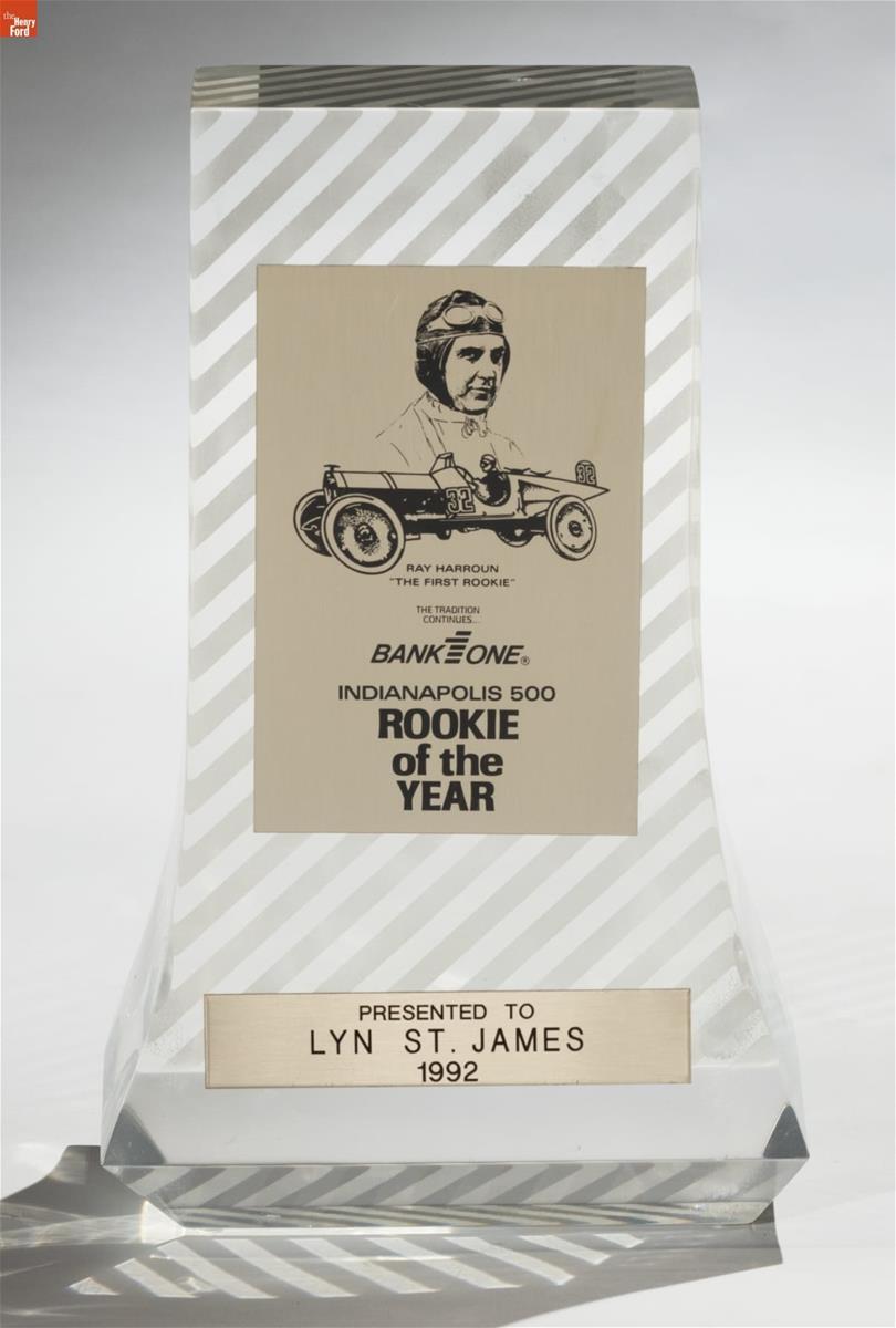 Clear glass or lucite trophy with diagonal stripes, and two panels with an image of a man in an old-fashioned racing helmet and goggles and an antique car, and text