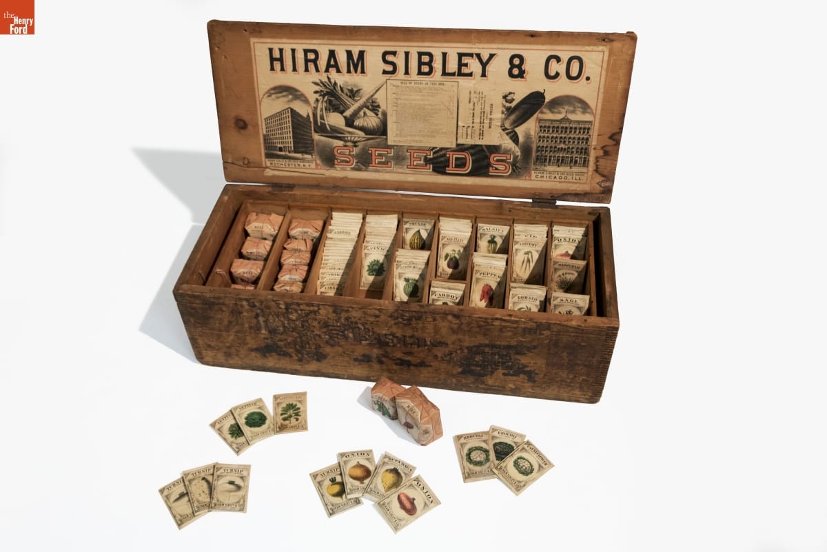 Open wooden box with graphics, images, and text on underside of lid; contains seed packets, with other seed packets arranged outside the box