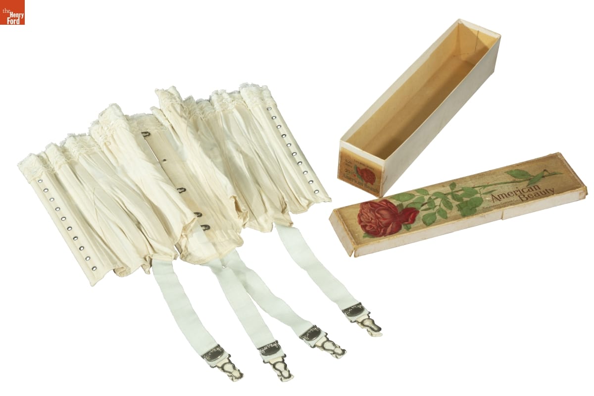 White corset with hook-and-eyes and belts with clips hanging down, next to box with image of red rose and text "American Beauty" 