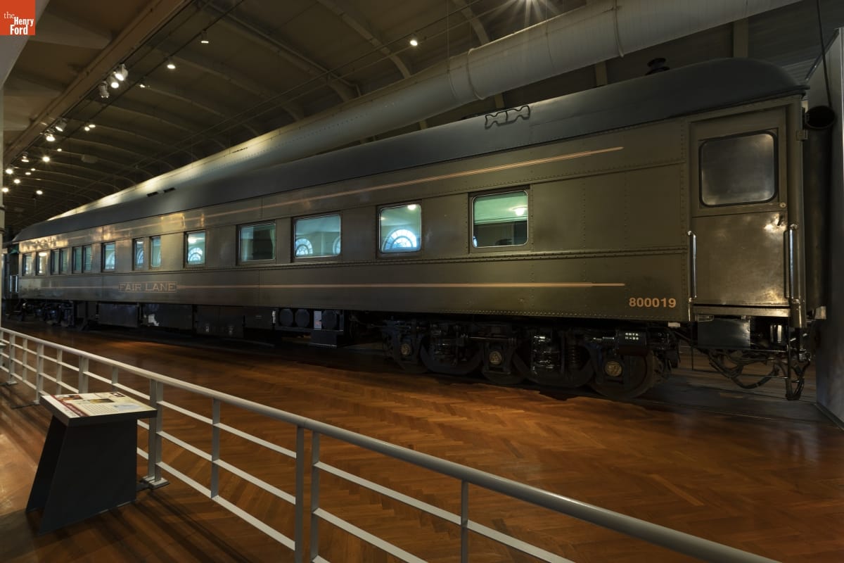 Long drab-green railcar, in large room with wood floor