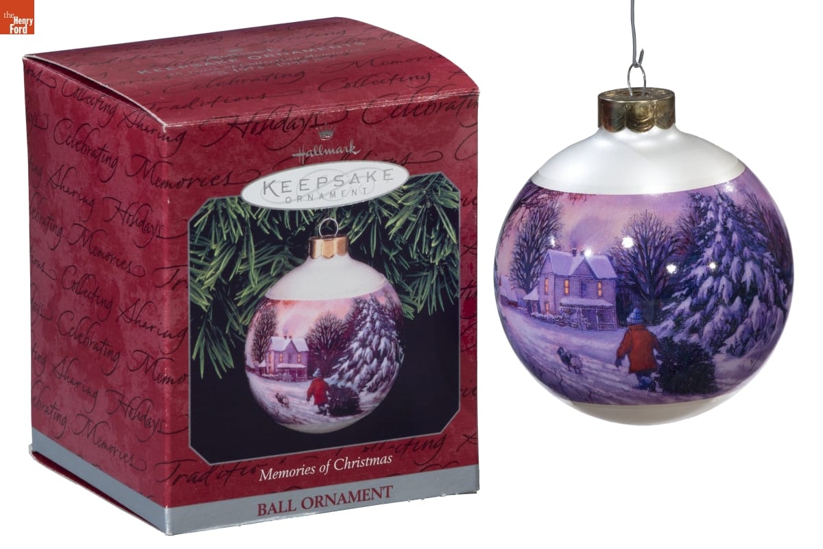 White bulb ornament with image of person dragging Christmas tree across a snowy field toward a house; box for ornament is also in photo