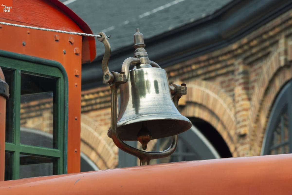 Locomotive Bell, 1927 Silver bell apparently mounted on top of orange locomotive (barely visible in picture) in front of a brick building