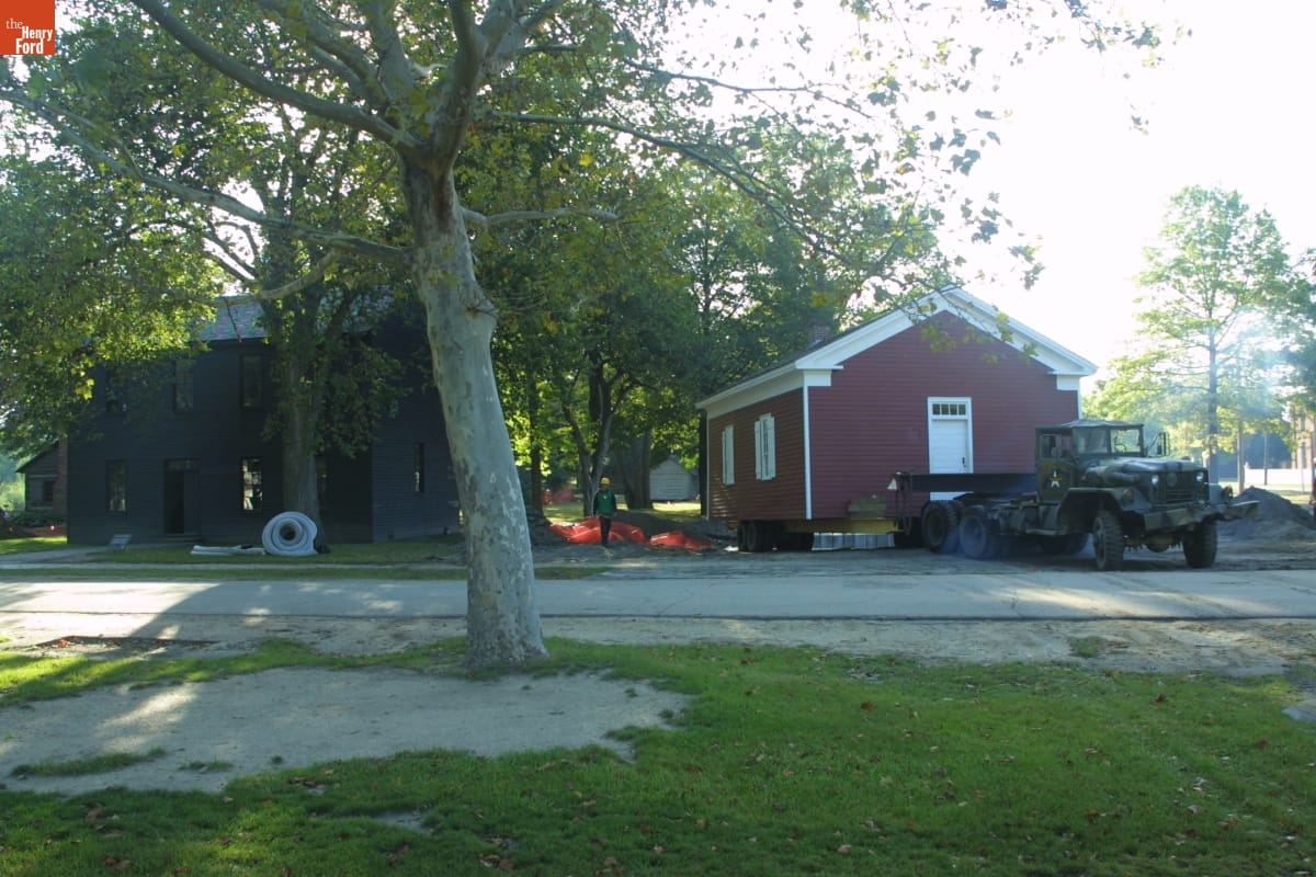 Small red wooden building on trailer behind truck next to gray wooden building among lawns, roads, and trees