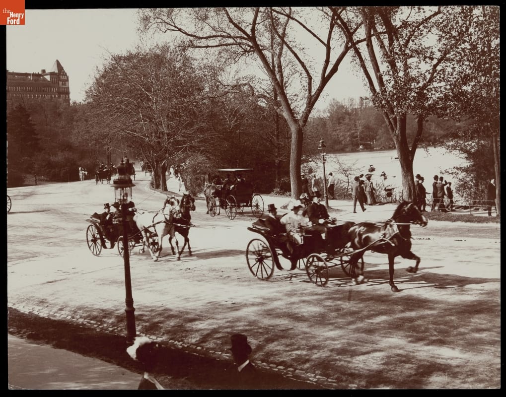 Horses pull carriages along a road or drive, with pedestrians strolling underneath trees by a lake nearby