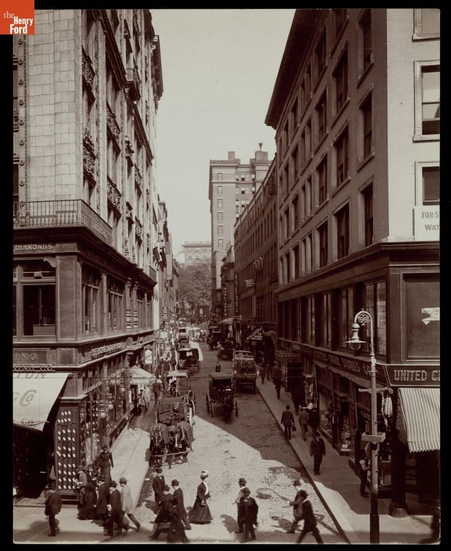 Street scene with pedestrians and carriages; tall buildings line both sides of street
