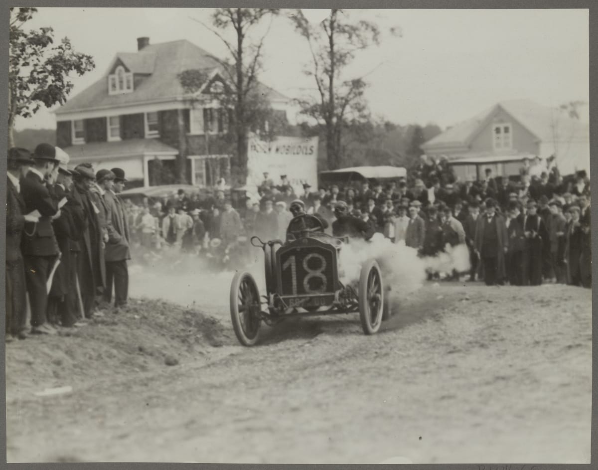 Lorraine-Detrich Automobile Driven by Arthur Duray at the Vanderbilt Cup Race, Long Island, New York, 1906 Early open race car careens around a corner on a dirt road, kicking up clouds of smoke with spectators looking on and houses in the background