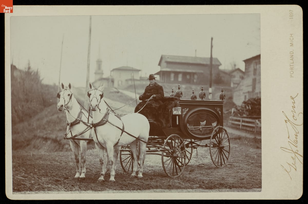 Low carriage with oval glass window and seated driver, drawn by two horses, stopped in front of a road and houses