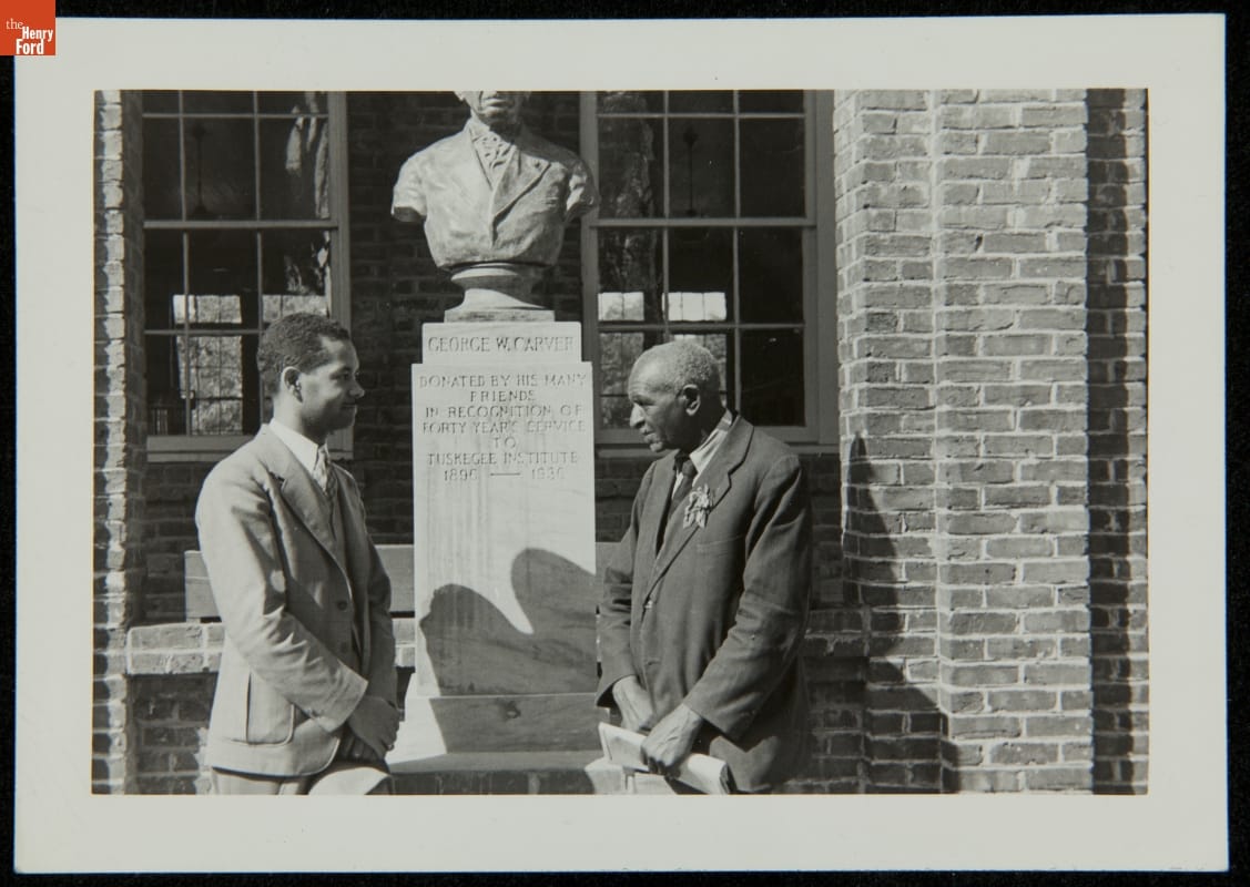George Washington Carver and Austin W. Curtis, Jr. at Tuskegee Institute with Sculpture by Steffen Thomas, circa 1938 Black-and-white photograph of two men in suits standing on either side of a sculpture of a man's head and torso above a tall base, all in front of a brick building with windows