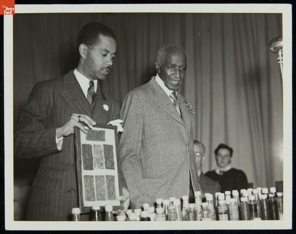 Black-and-white photo of two men in suits standing at table filled with bottles; one man holds a small display board