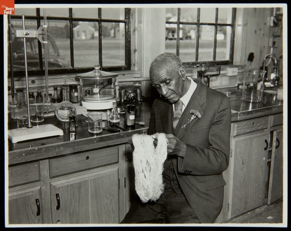 Black man in suit with flower in boutonnière sits by a counter full of scientific equipment and examines yarn or fiber