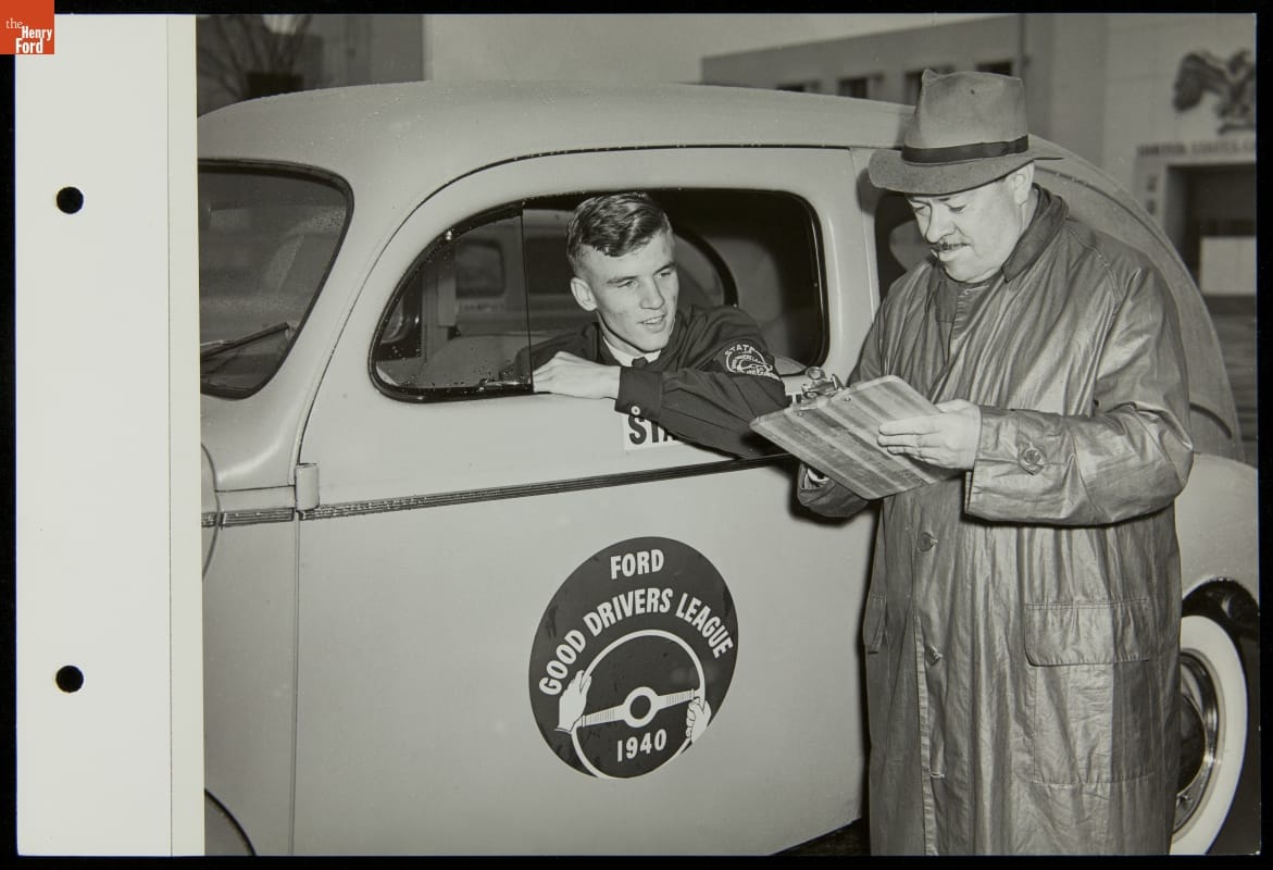 Young Man Taking Driver's Test, Ford Good Drivers League, Ford Exposition, New York World's Fair, 1940 Man in raincoat and hat holds a paper by a car with decal with text on side; young man sticks head and elbow out driver side window