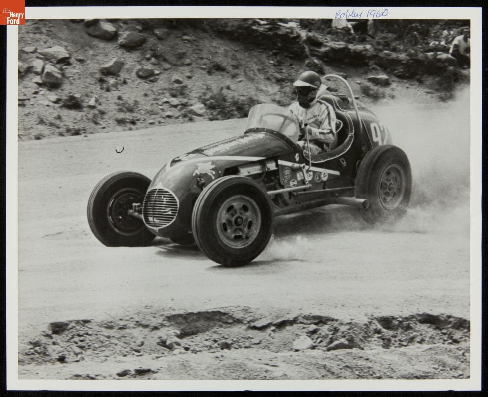 Bobby Unser, Racing at Pikes Peak, 1960 Person wearing face mask, helmet, and jumpsuit drives race car on dirt road with rocky slopes on either side