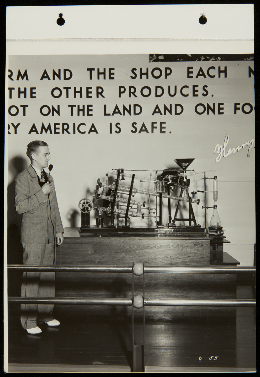 Presenting the Soybean Extractor, Texas Centennial Central Exposition, Dallas, Texas, 1936 Person in suit holding microphone stands next to a piece of equipment under text on a wall