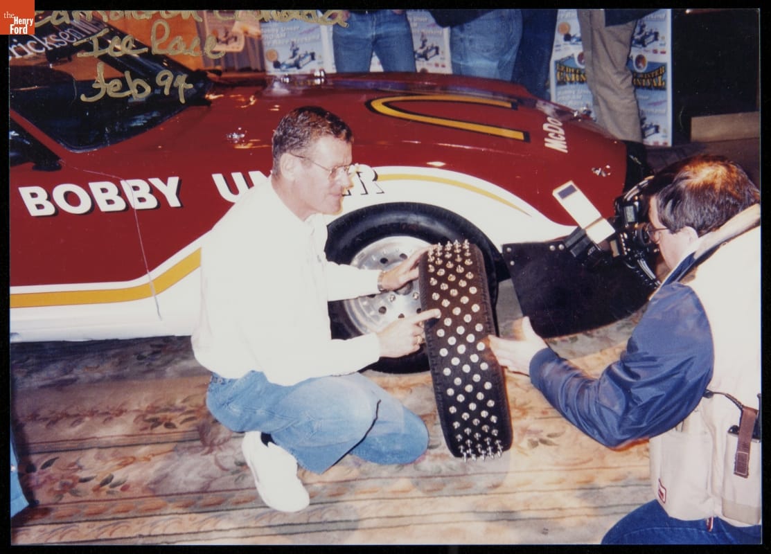 Man kneels next to a car pointing to a studded tire he is holding, while another man kneels nearby