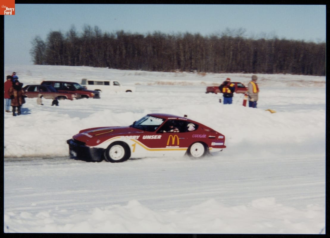 Bobby Unser ice racing in Canada, 1994 Red and white race car on a snowy road with snowbanks on either side, with people, other cars, and trees in background