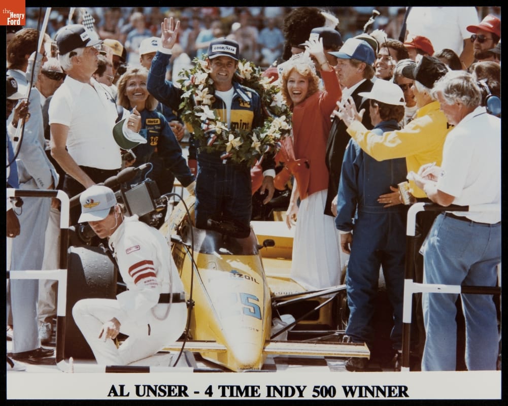Man in jumpsuit with wreath around neck stands in a race car waving to the camera with a crowd of people looking on