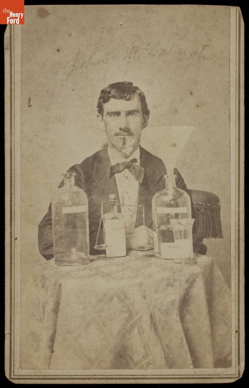 Black-and-white portrait of man in suit sitting at table containing bottles and jars