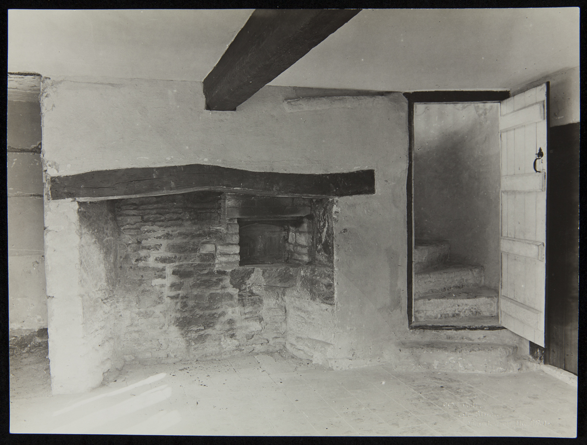 Interior of Cotswold Cottage at its Original Site in Chedworth, Gloucestershire, England, 1929-1930 Bare interior of room with low ceiling, large fireplace, and door open to reveal spiral staircase leading upwards