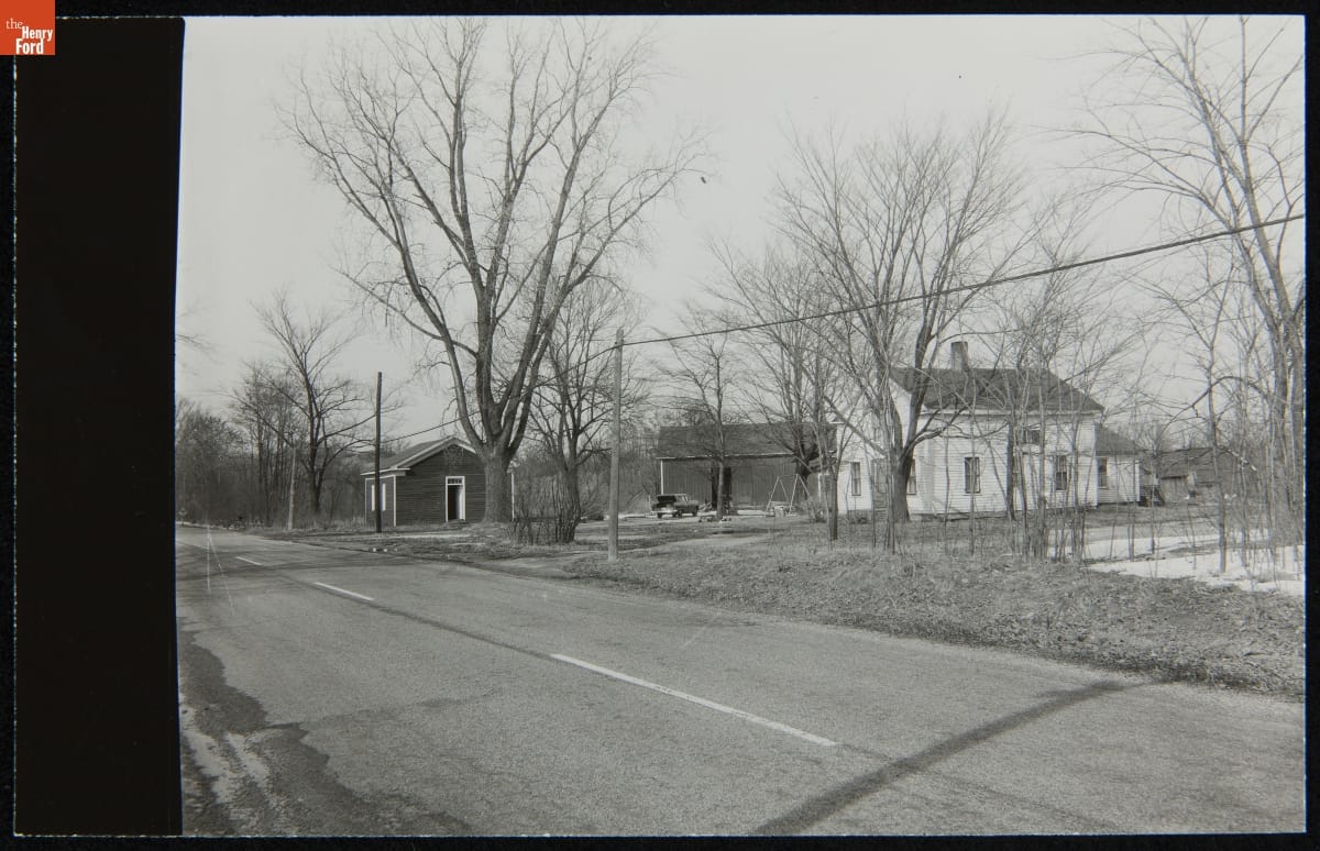 Black-and-white photo of several wooden buildings by side of two-lane road among trees