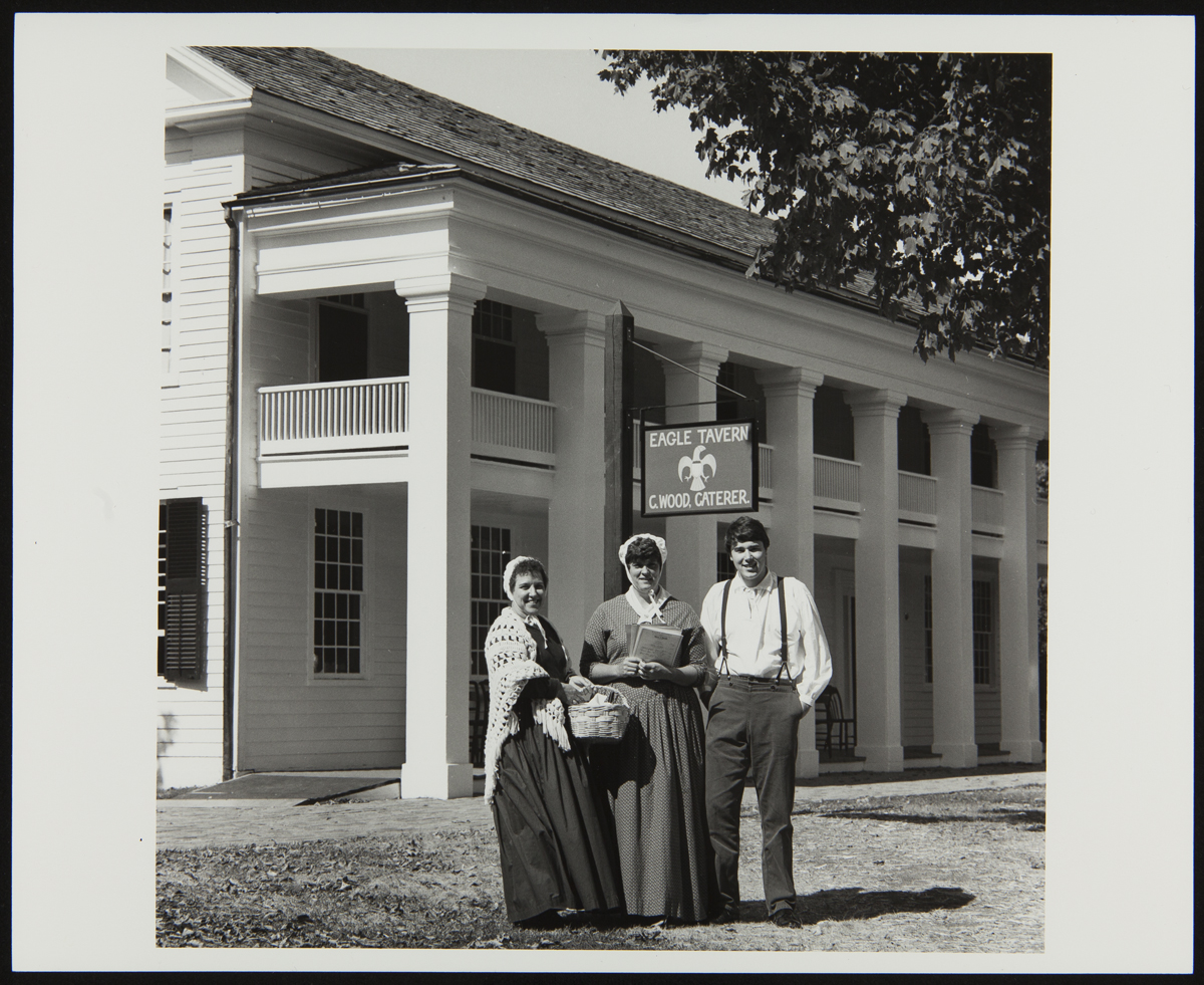 Three people in historical garments stand outside a two-story wooden building with sign containing text 