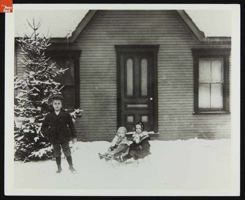 Two children sit on a sleigh in snow in front of a door; another boy stands nearby in front of a tree