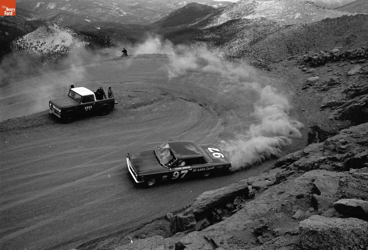 Car rounding an uphill hairpin curve in a cloud of dust on a dirt road with mountains in the background