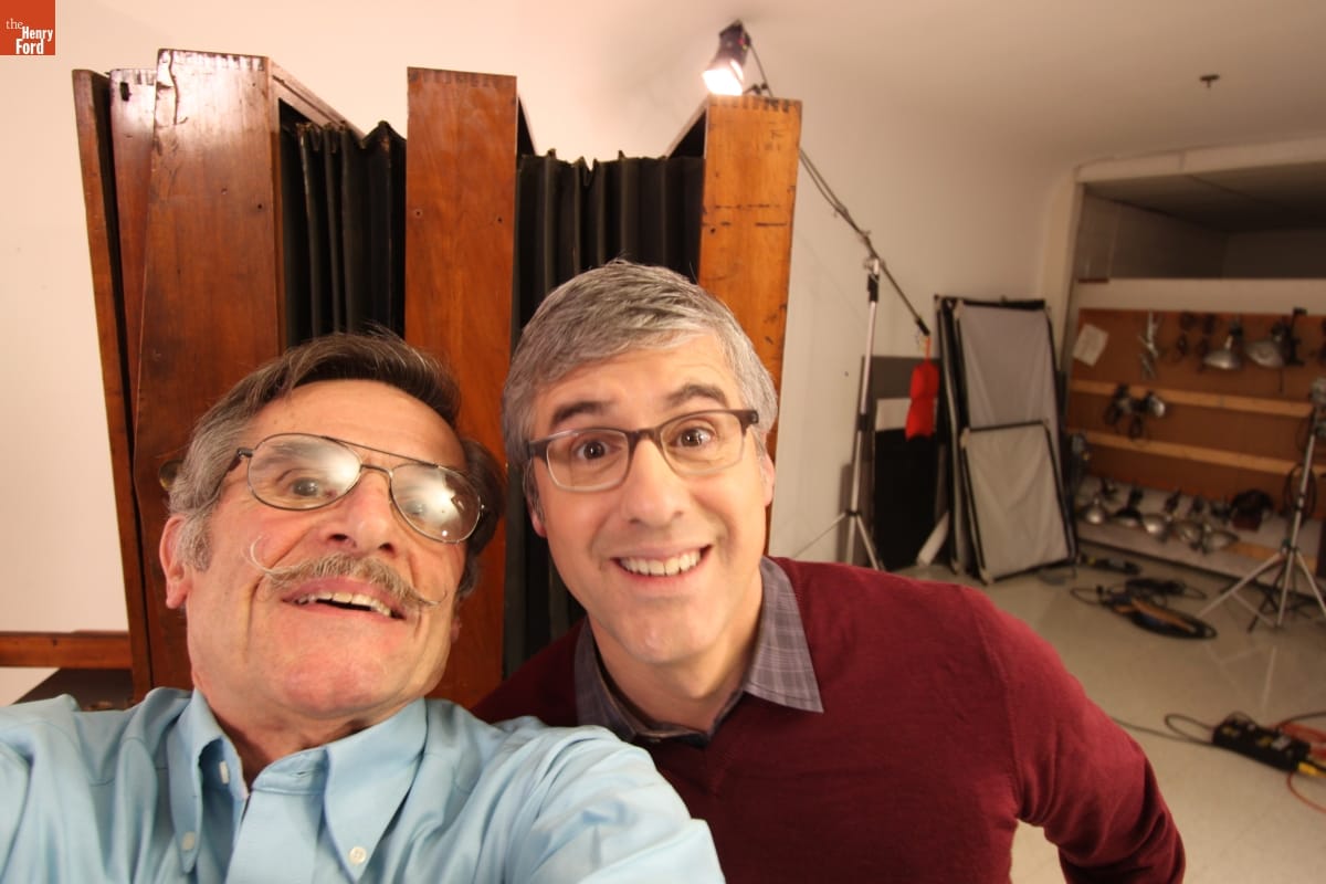 "Selfie" of Rudy Ruzicska and Mo Rocca in the Henry Ford Museum Photographic Studio, December 11, 2015 Two men smile smile for a selfie with a large old-fashioned box camera and modern photography equipment behind them