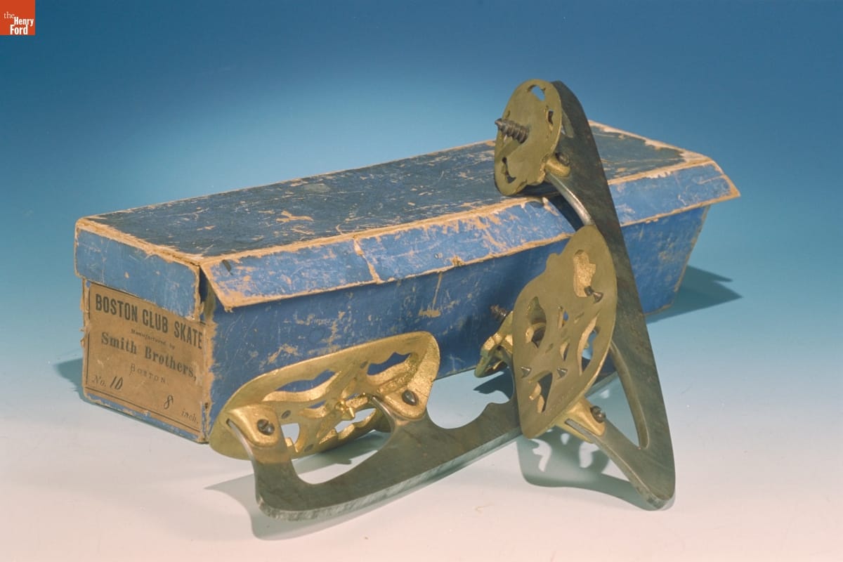 Ice Skates, 1866-1870 Blue shoebox with tan label on end and two gold and silver ice skating blades next to it