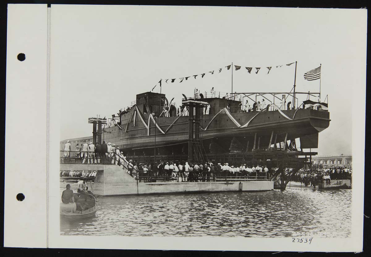 Eagle Boat #60 Lowered to Water, August 1919 Long, narrow boat decorated with flags and bunting on elevated trestle above water, as a crowd looks on