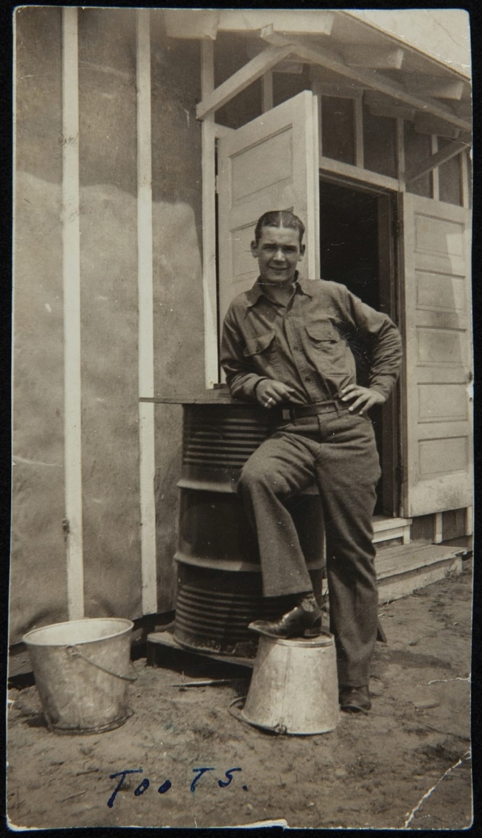 Man wearing uniform leans on piece of equipment with one foot on upturned bucket outside structure