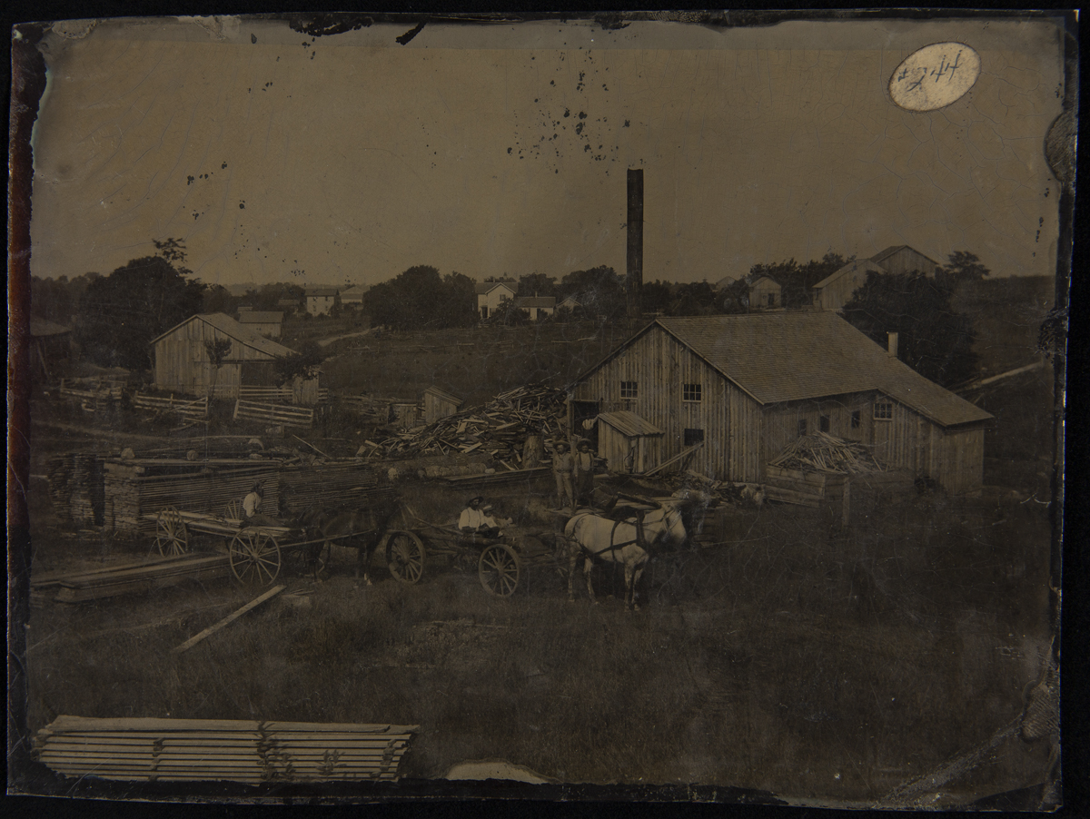 Landscape with wooden buildings, stacks and piles of lumber, horses and wagons, and people