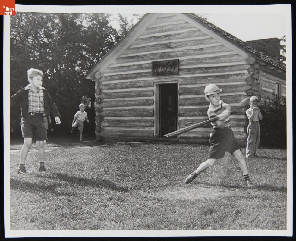 Four boys stand in front of a log cabin; one swings a bat