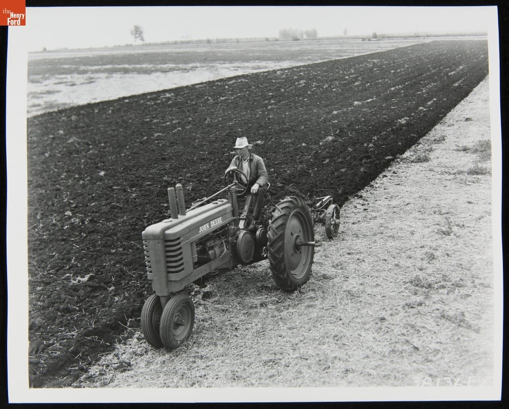 Black-and-white photo of man riding a tractor through a field partially dirt and partially stubble