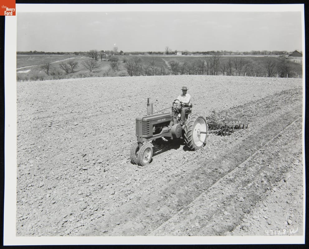 Black-and-white photo of man riding a tractor through a dirt field