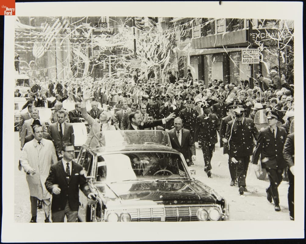 Parade with people standing in an open car, waving; uniformed officers walking alongside; and confetti and tickertape in the air