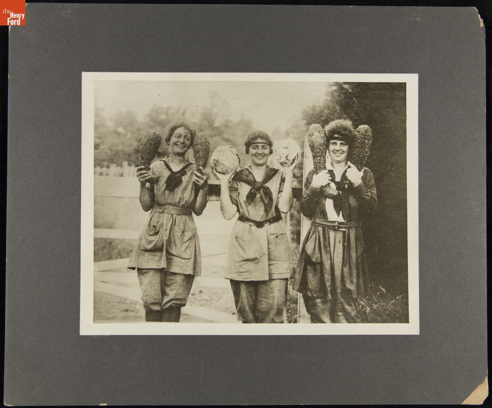 Members of the Woman's National Farm and Garden Association, 1918 Black-and-white image of women, perhaps in some type of uniform, holding vegetables up in each hand