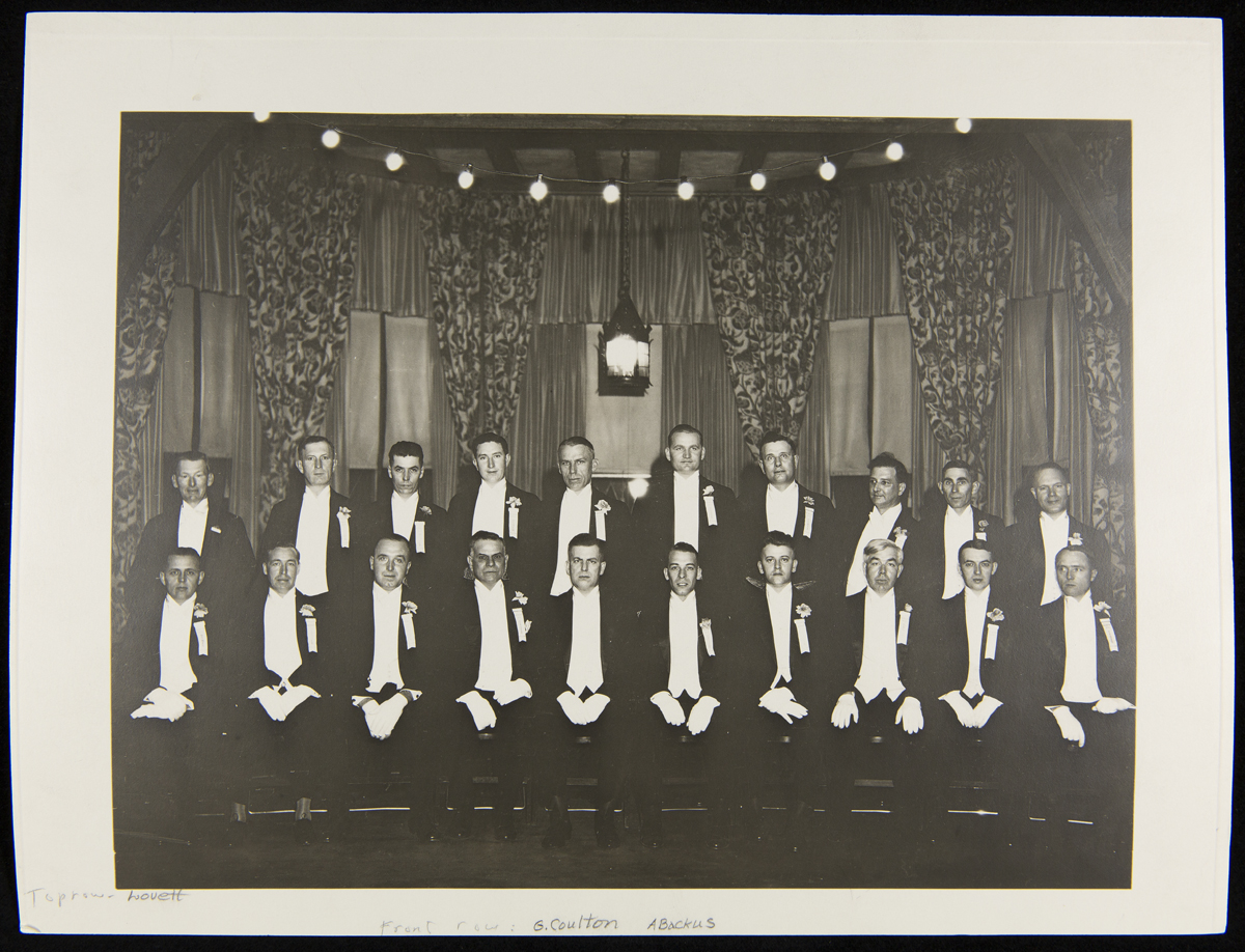 Men in tuxedos and white gloves pose for a photo, some standing and some sitting