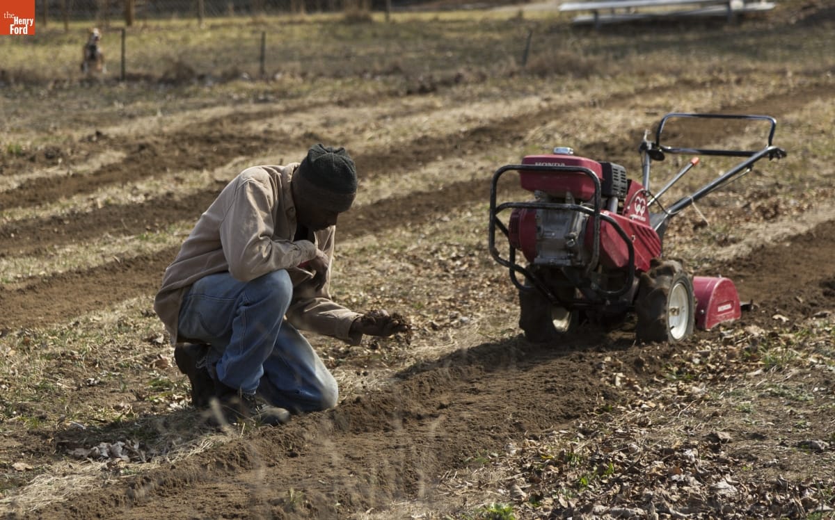 Melvin Parson Gardening during the Entrepreneurship Interview A man kneels in a field by a piece of equipment, looking at a handful of dirt he is holding