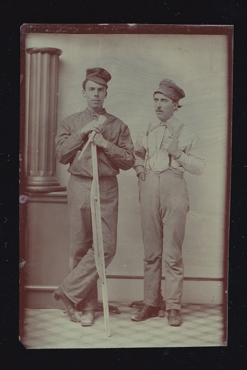 Two men in caps, one holding a long tool, standing next to a pillar