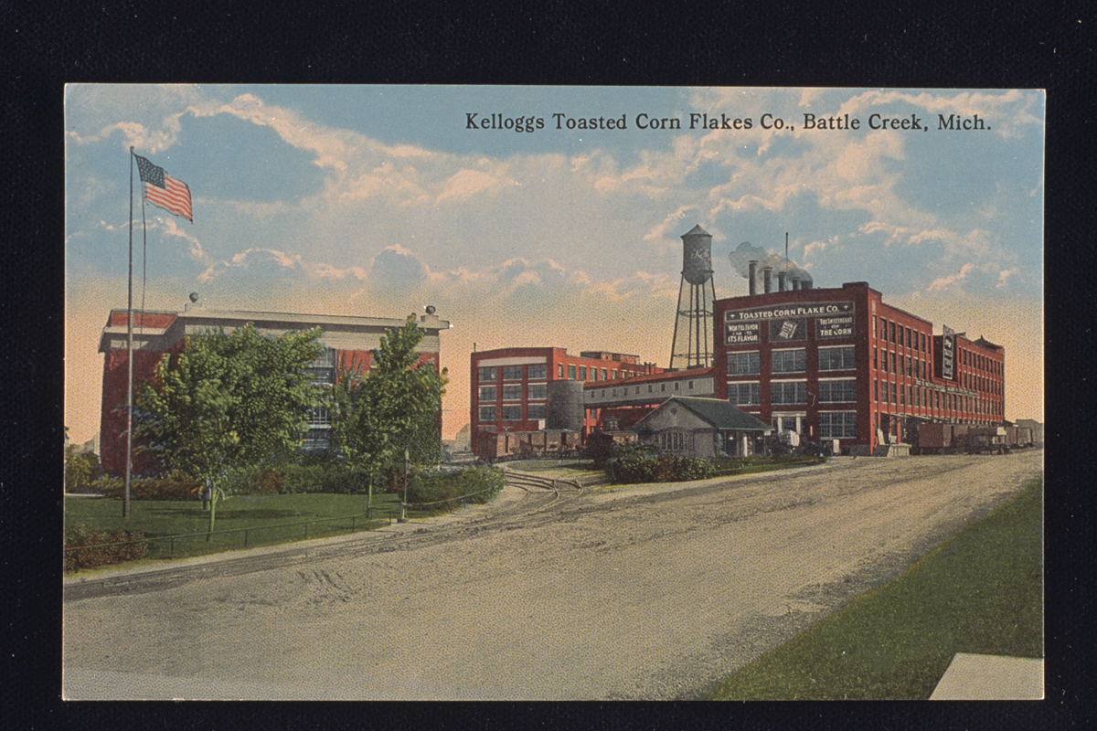 Postcard of Kellogg’s Toasted Corn Flake Company factory, 1914, showing several red brick buildings, a water tower, and an American flag in the foreground