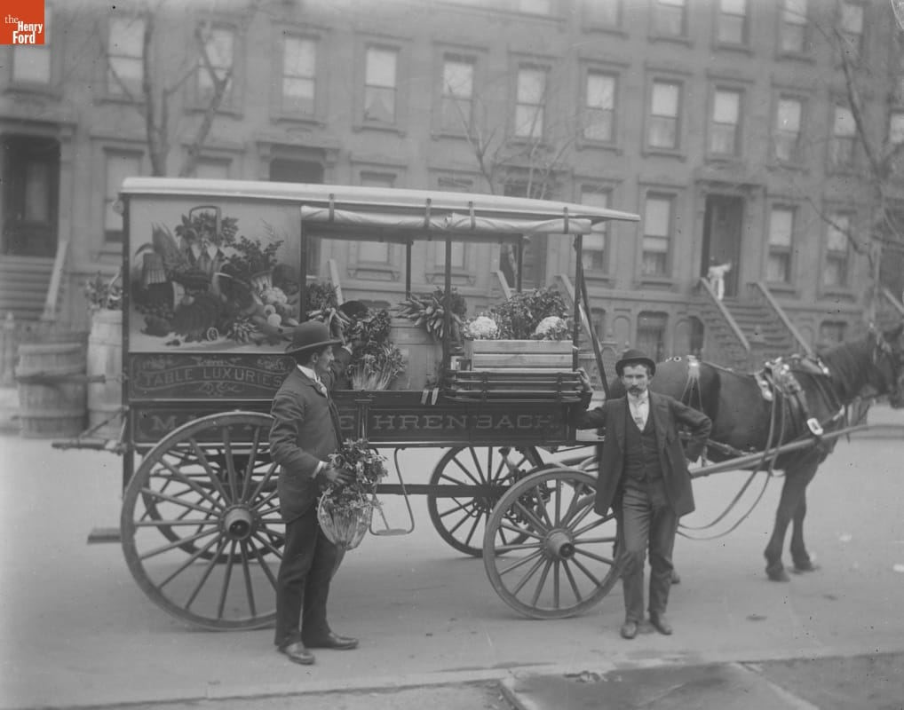 Two men stand by a horse-drawn wagon, decorated with a painting of fruits and vegetables and containing produce