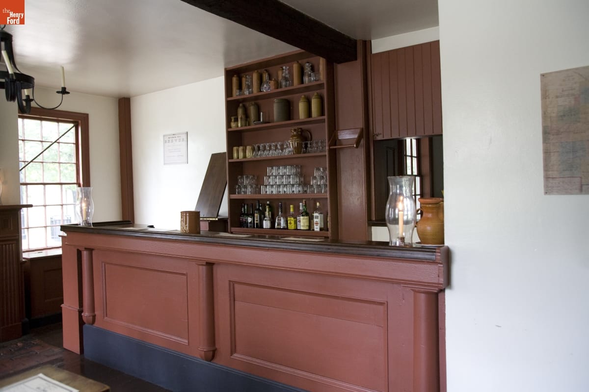 Room with a brown wooden bar, behind which are shelves containing liquor bottles, glasses, crocks, and decanters