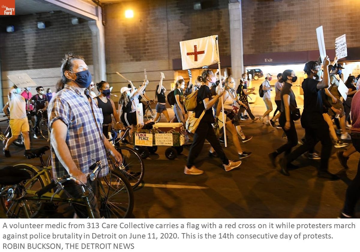 Photo of people in protest march, some walking bicycles or holding signs; person in center carries a white flag with a red cross; also contains text
