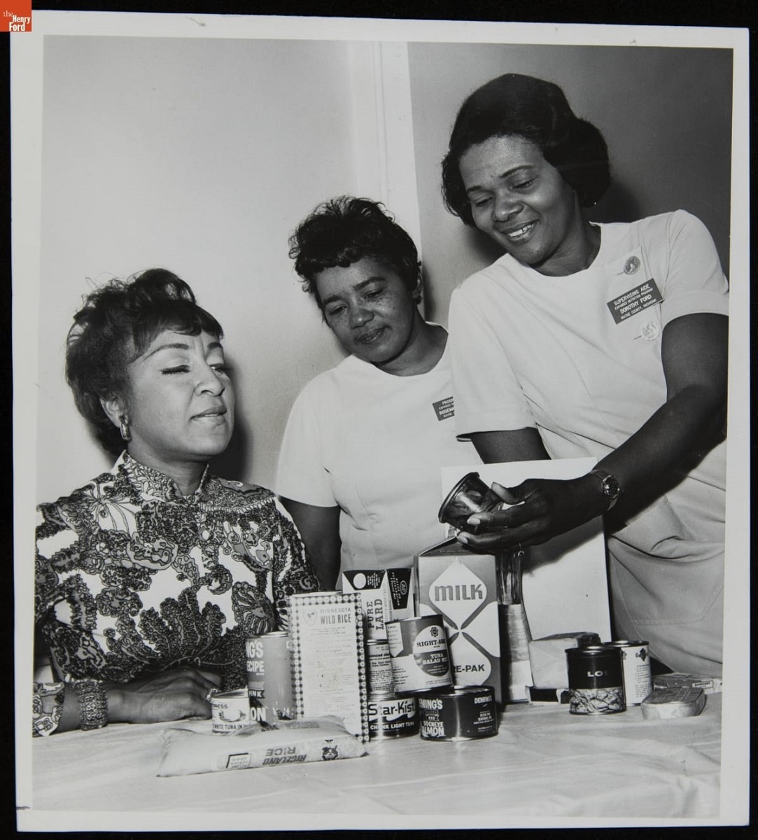 June Sears, Rosemary Dishman, and Dorothy Ford Discussing Women's Nutrition, May 1970 Three Black women sit and stand around a table that holds food packages