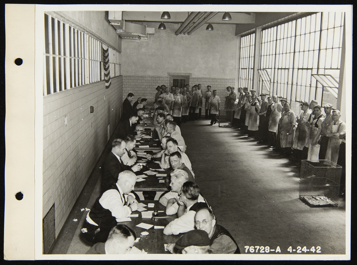 Large room with small tables lined up along one side, each with people sitting on either side, and a line of people snaking around the other side of the room 