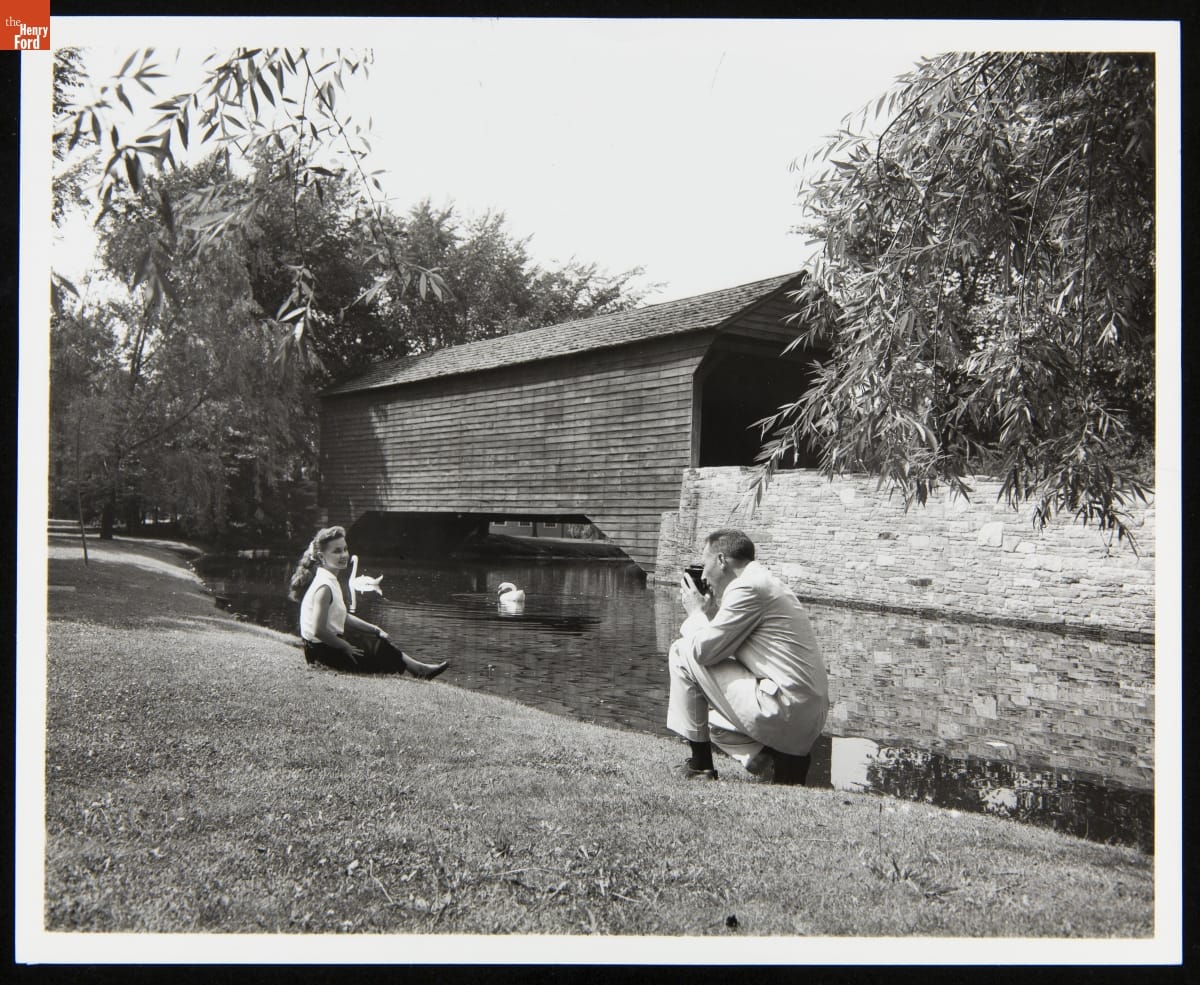Black-and-white photograph of man, kneeling, photographing woman seated on grass, with pond, swans, and a wooden covered bridge behind them