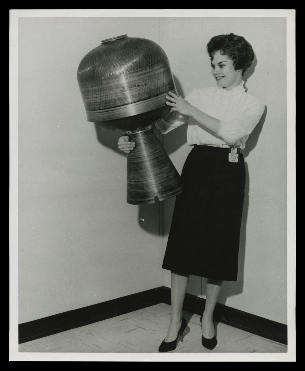 Beverly Rushton Holding Up a Retrorocket for NASA Ranger Spacecraft, 1962 Black-and-white photo of smiling woman holding contraption with cone-shaped bottom section and dome-shaped top section