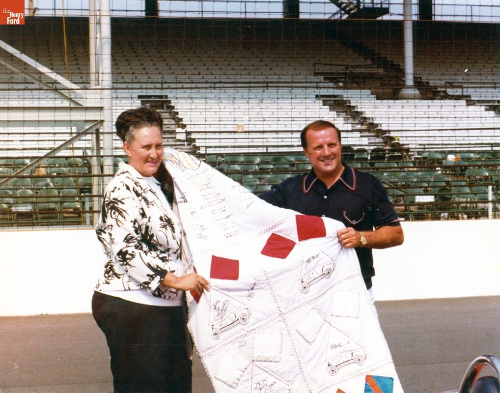 Jeanetta Holder Presents A.J. Foyt with a Quilt as the Winner of the 1977 Indianapolis 500 Woman with dark hair in braid wrapped around her head and man in polo shirt hold a quilt in front of empty grandstands